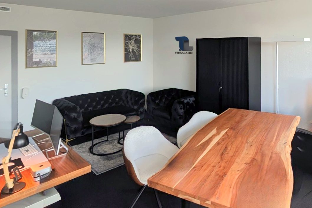 Le bureau de la direction, coin réunion : table en bois massif avec deux fauteuils blancs, un canapé et un fauteuil noirs capitonnés autour de deux tables basses rondes, trois cadres et le logo TL Fiduciaire au mur, une armoire noire à droite.